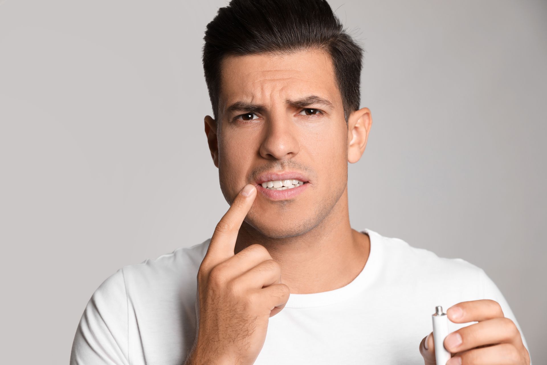 Man in white shirt touching a front tooth, holding a small dental item, with a pained expression against a gray background