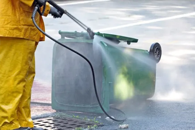 Person pressure-washing a tipped-over green trash bin on pavement, with water spraying around it