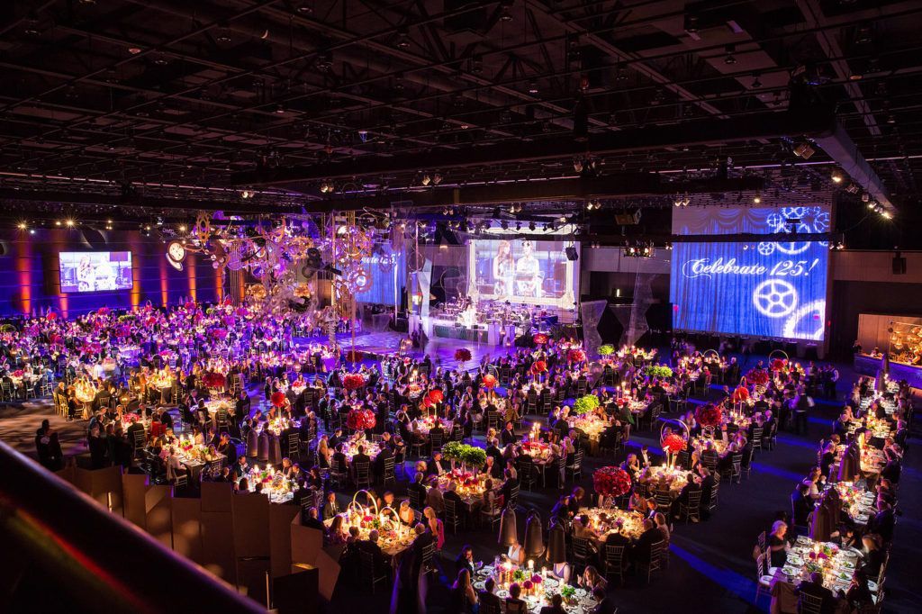 Large banquet hall filled with round tables and colorful stage lighting at a formal event
