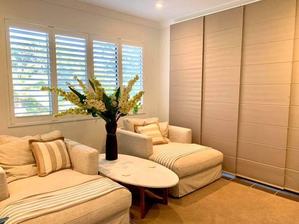 Cozy beige seating area with window shutters and panel blinds; vase of flowers on a table.