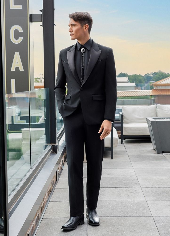 Man in black suit and bolo tie, standing on a rooftop, looking to the side.