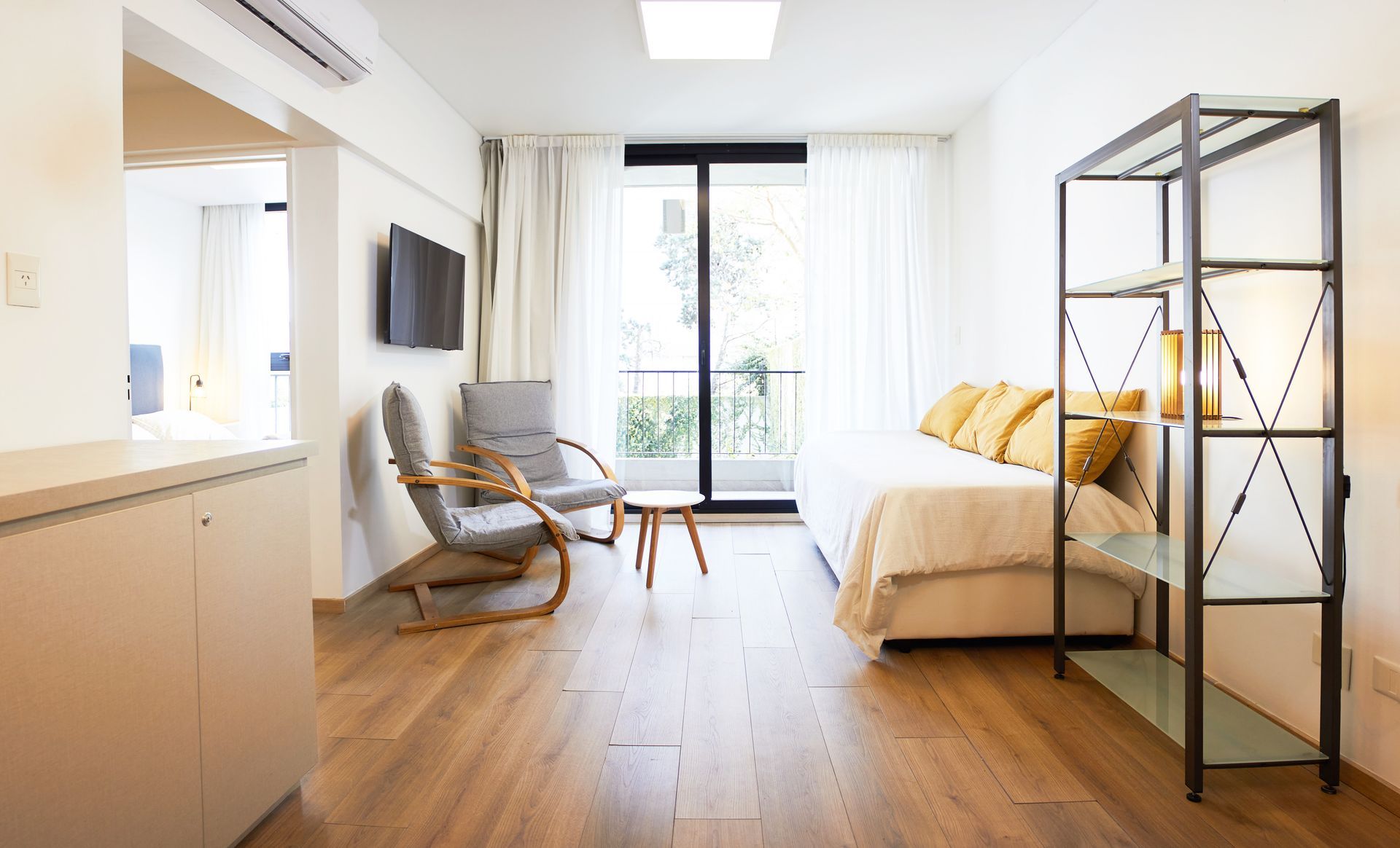 Estudio luminoso y minimalista con cama, silla, estanterías y grandes ventanales que dejan entrar la luz del sol.
