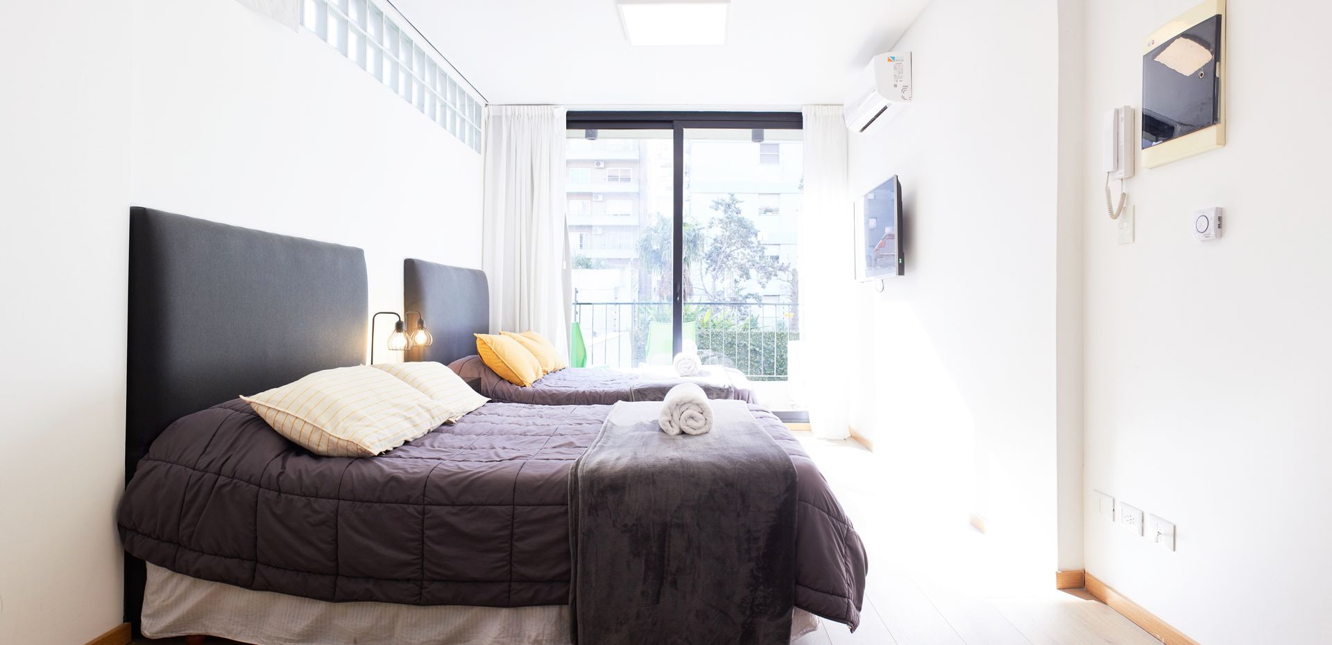 Habitación luminosa con cama, almohadas y ventana grande en una habitación blanca minimalista.