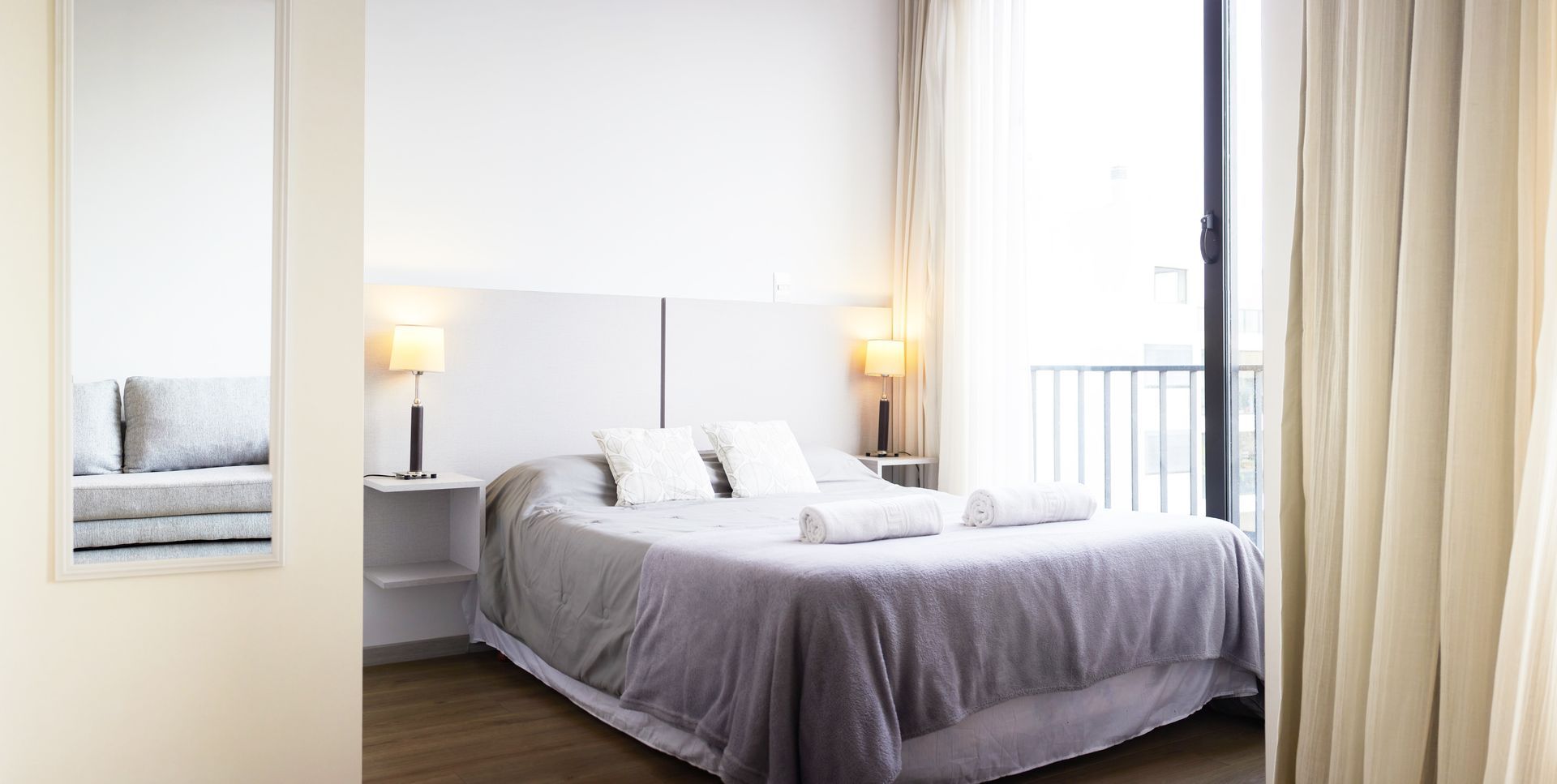 Habitación luminosa con una cama bien hecha, lámpara de noche y un gran espejo junto a la ventana.