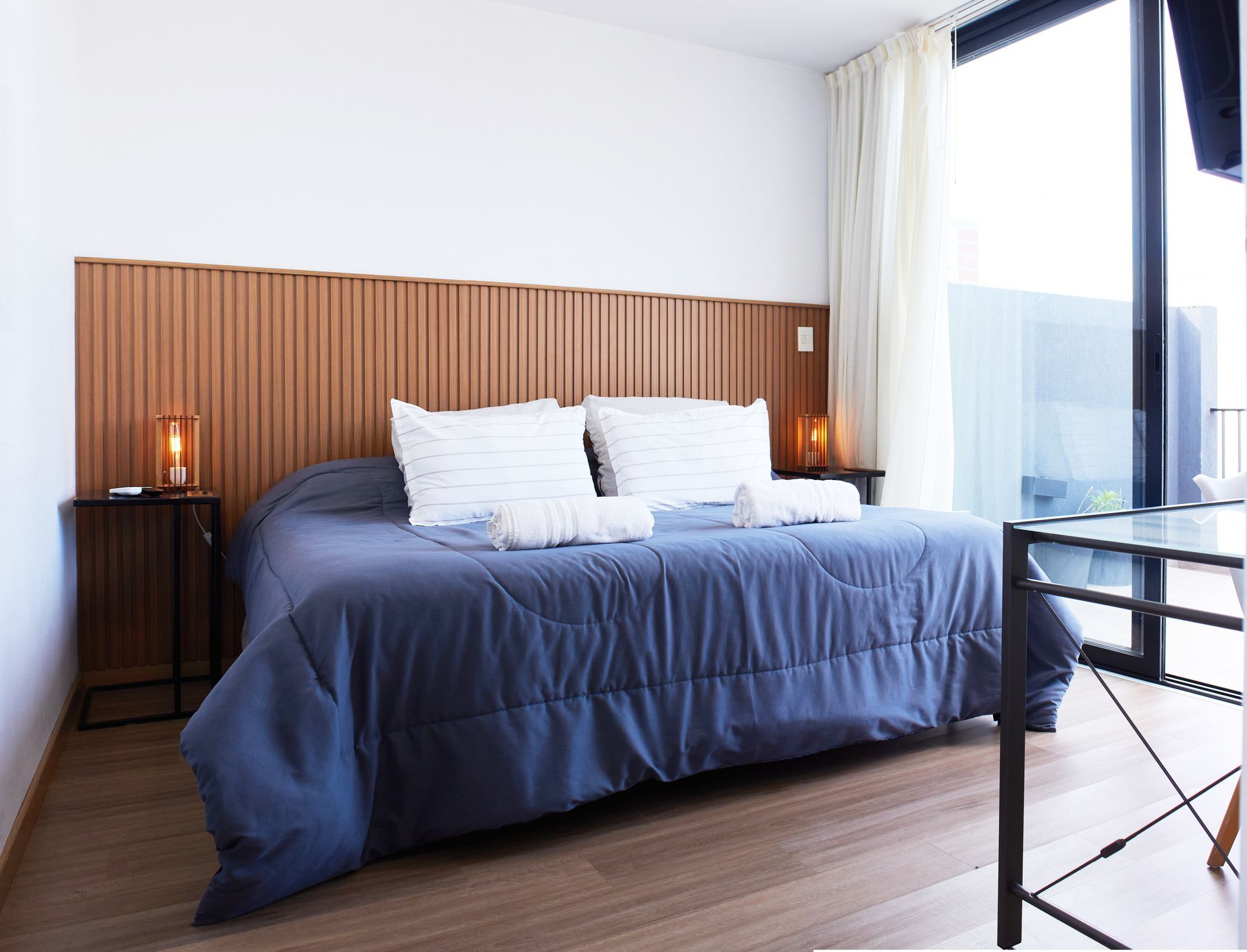 Dormitorio moderno con colcha azul marino, cabecero de madera, lámparas de noche y ventanal de suelo a techo.