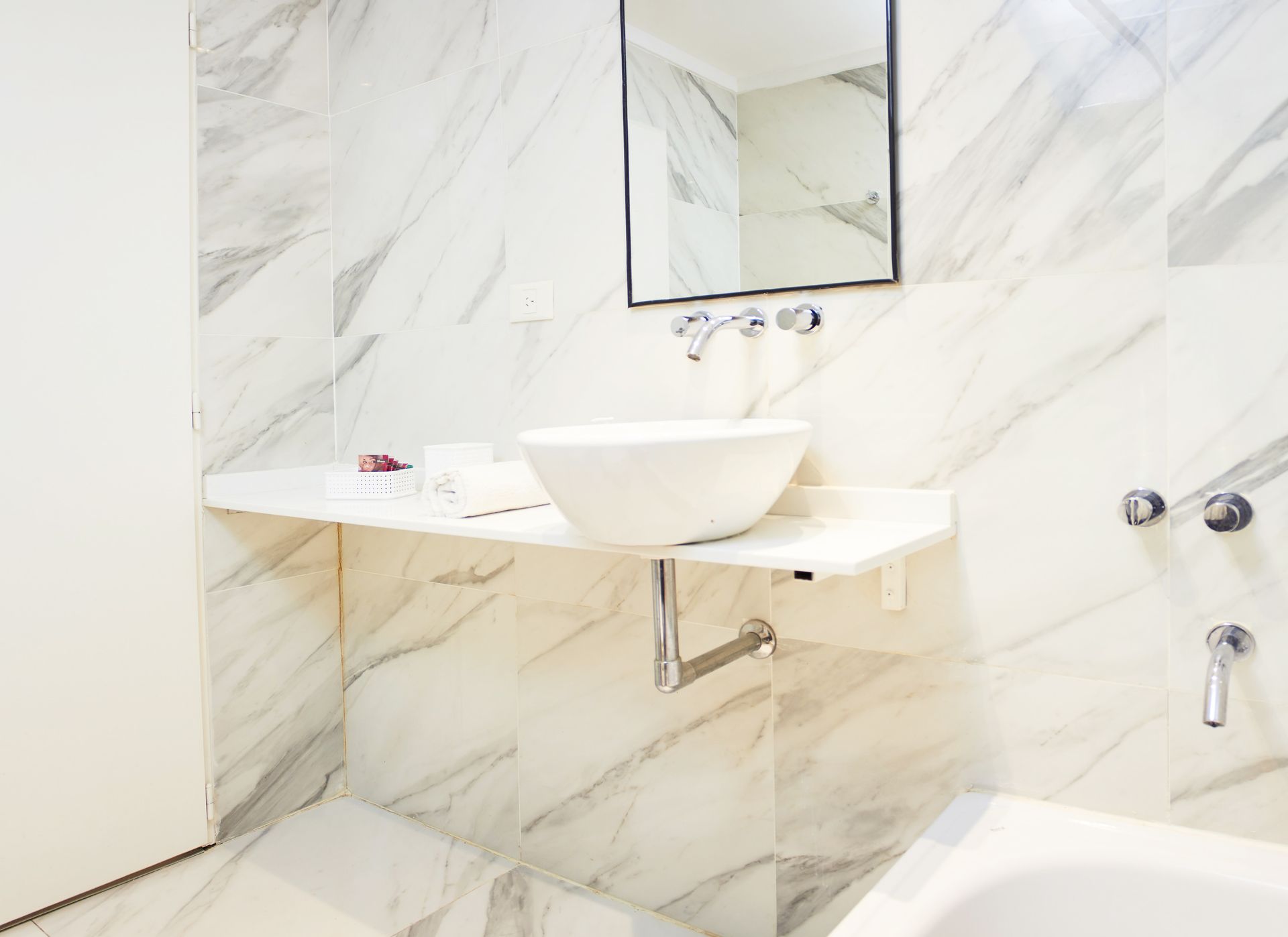 Baño moderno con paredes de mármol blanco, lavabo mural, espejo y grifería para bañera.