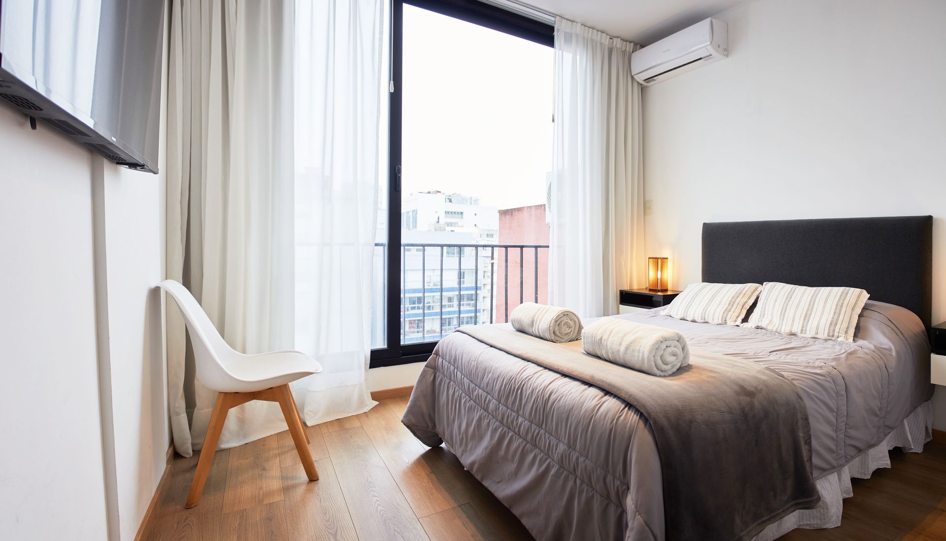 Habitación luminosa con cama, silla, lámpara y una gran ventana con cortinas y puerta de balcón.
