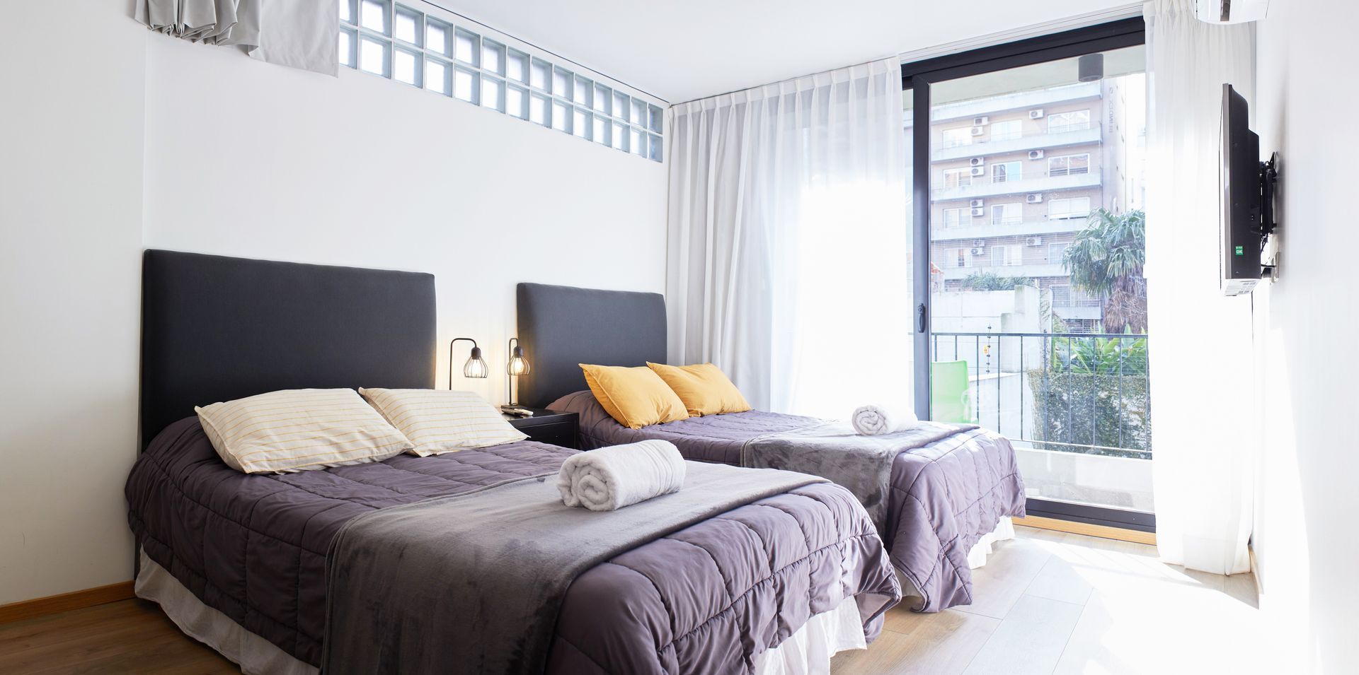 Habitación luminosa con dos camas, paredes blancas y una gran ventana con cortinas transparentes.