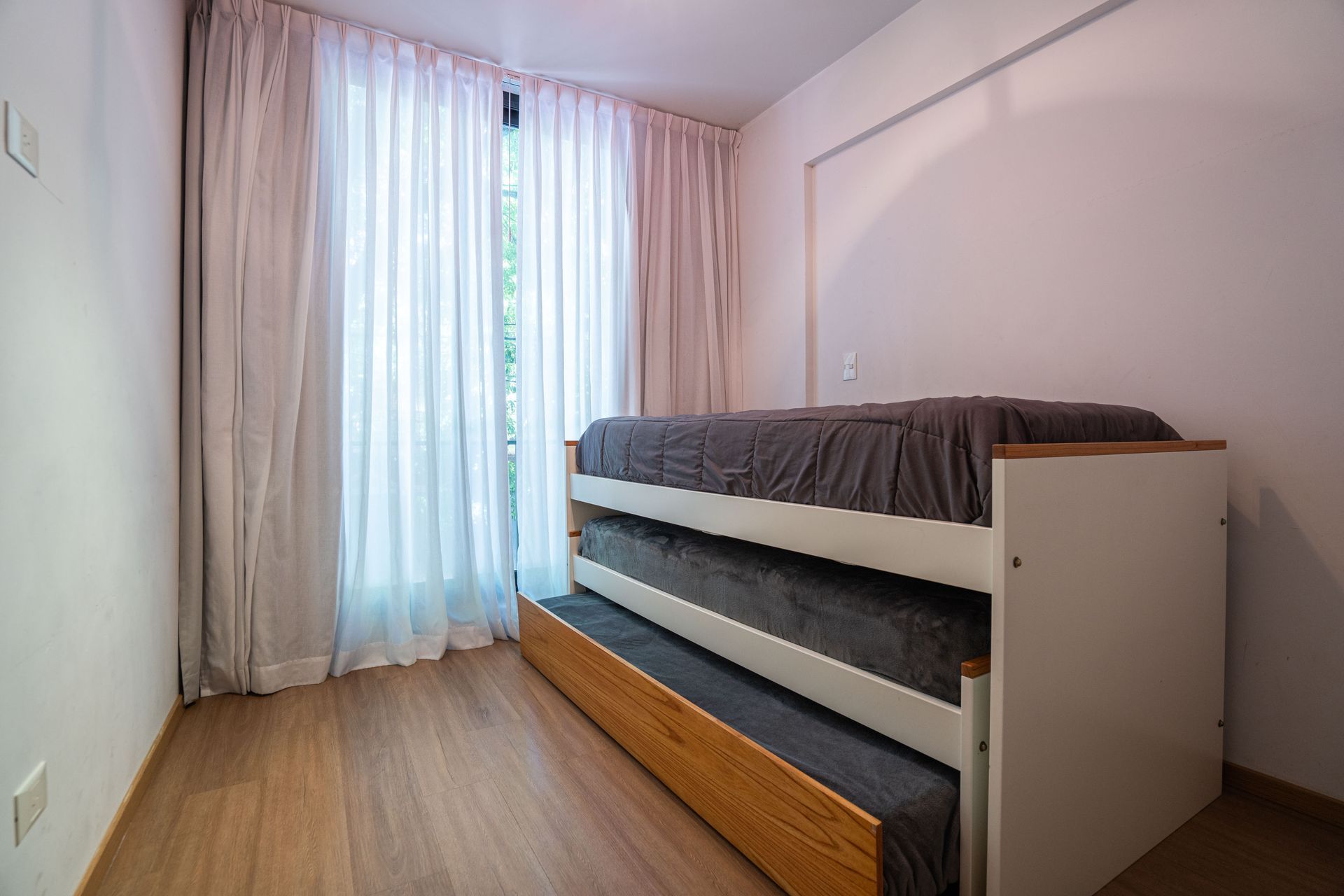 Habitación minimalista con dos camas individuales, suelo de madera y cortinas transparentes junto a una ventana luminosa.