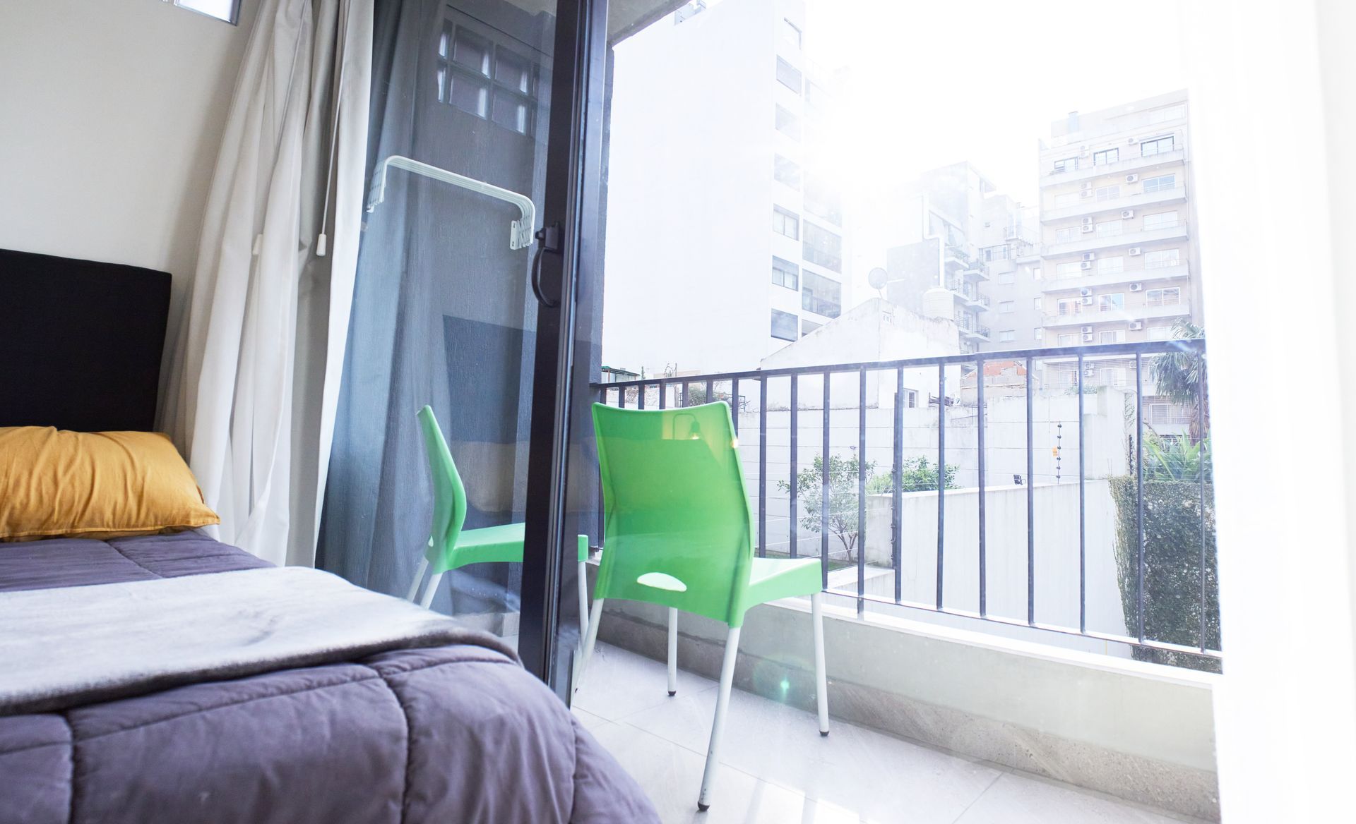 Balcón luminoso en el dormitorio con sillas verdes, barandilla de metal y una cama junto a cortinas transparentes.