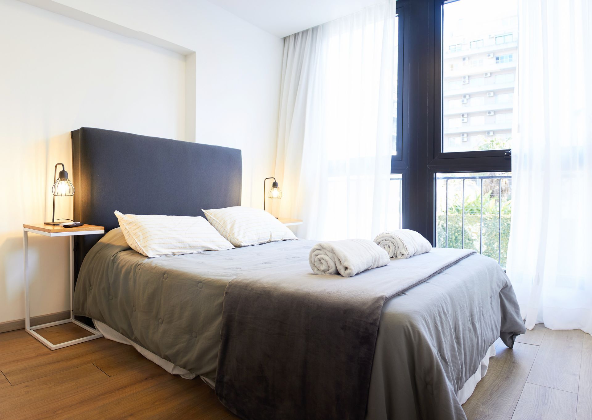 Habitación luminosa con una colcha gris, sábanas blancas, lámparas de noche y grandes ventanas con cortinas transparentes.