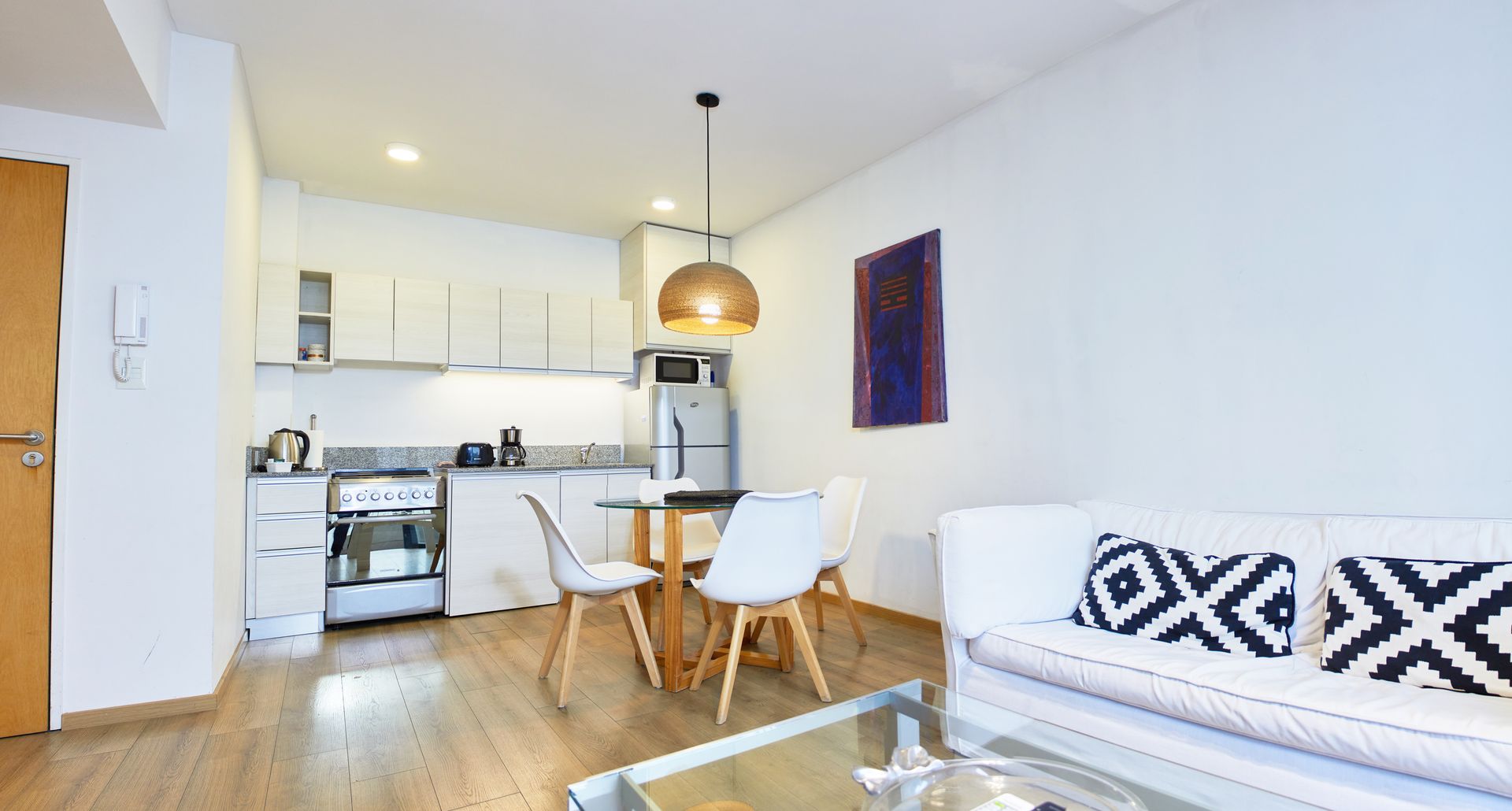 Apartamento tipo estudio luminoso y diáfano con cocina, mesa de comedor, sofá blanco y suelos de madera.
