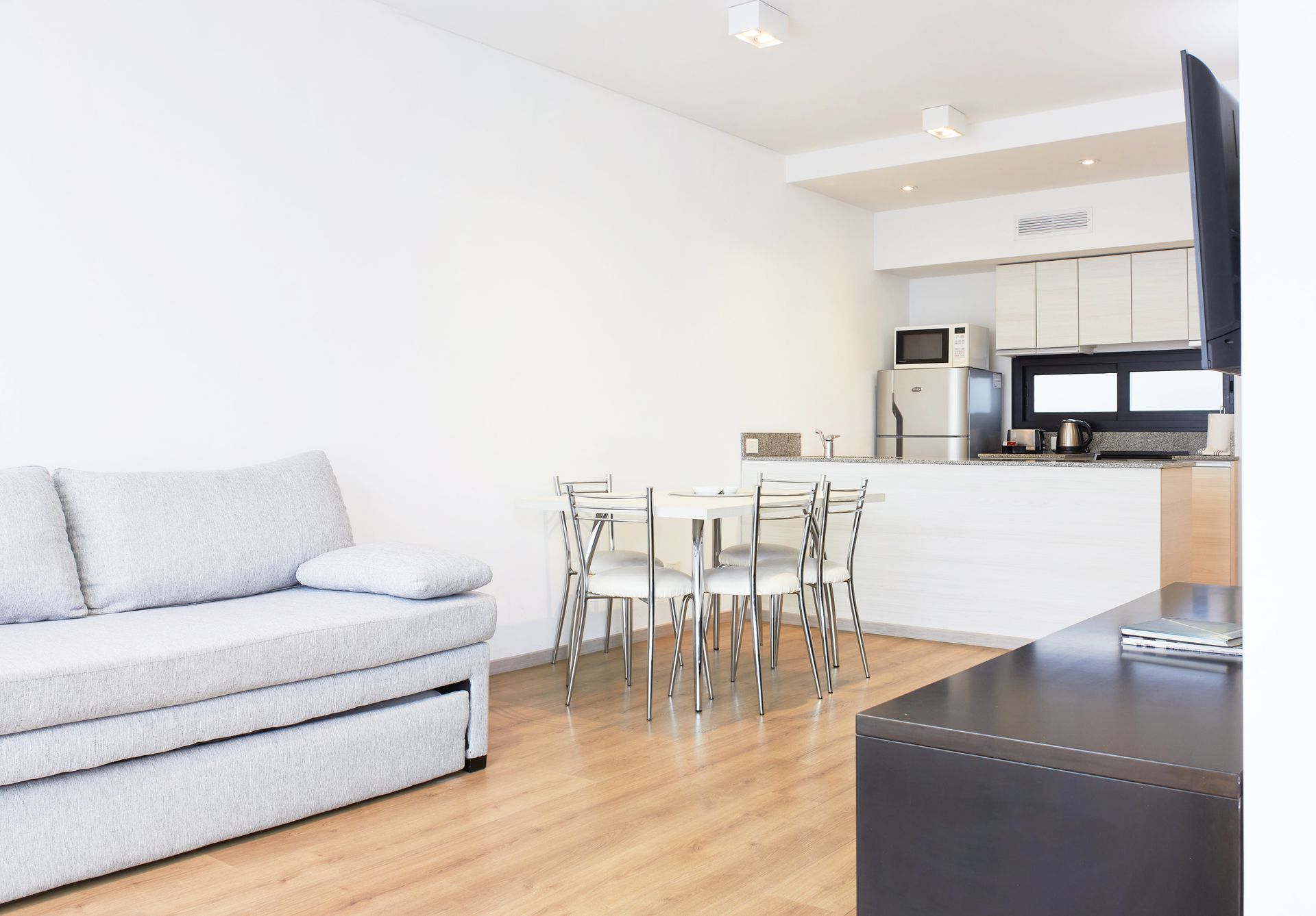 Luminoso salón moderno con sofá gris, mesa de comedor de cristal y zona de cocina blanca con suelos de madera.