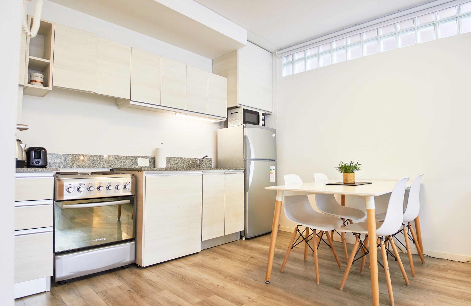 Luminosa zona de cocina-comedor con armarios blancos, suelo de madera, una mesa y un frigorífico de acero inoxidable.