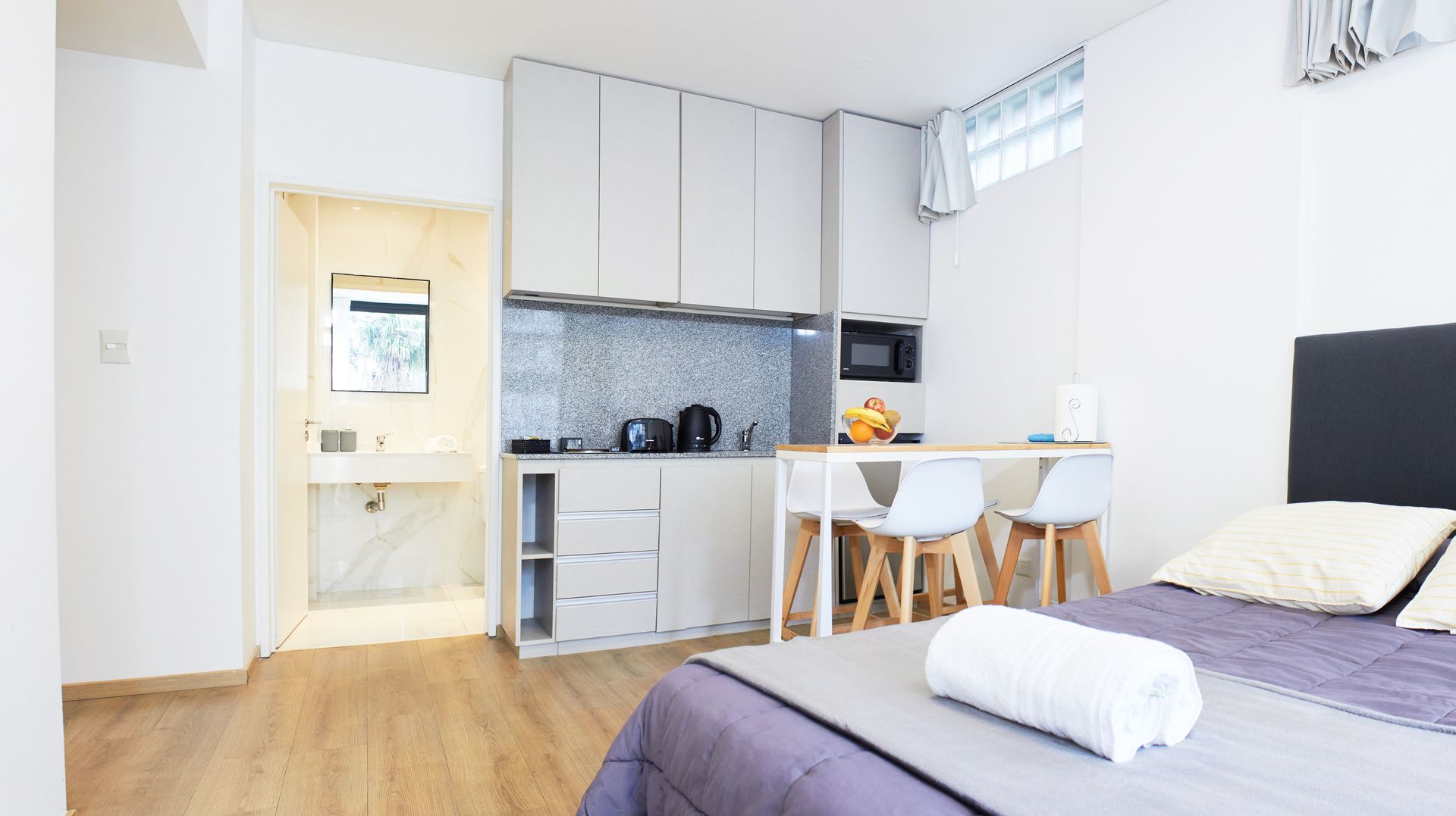 Apartamento tipo estudio luminoso con cama, cocina americana, mesa de comedor y decoración blanca.