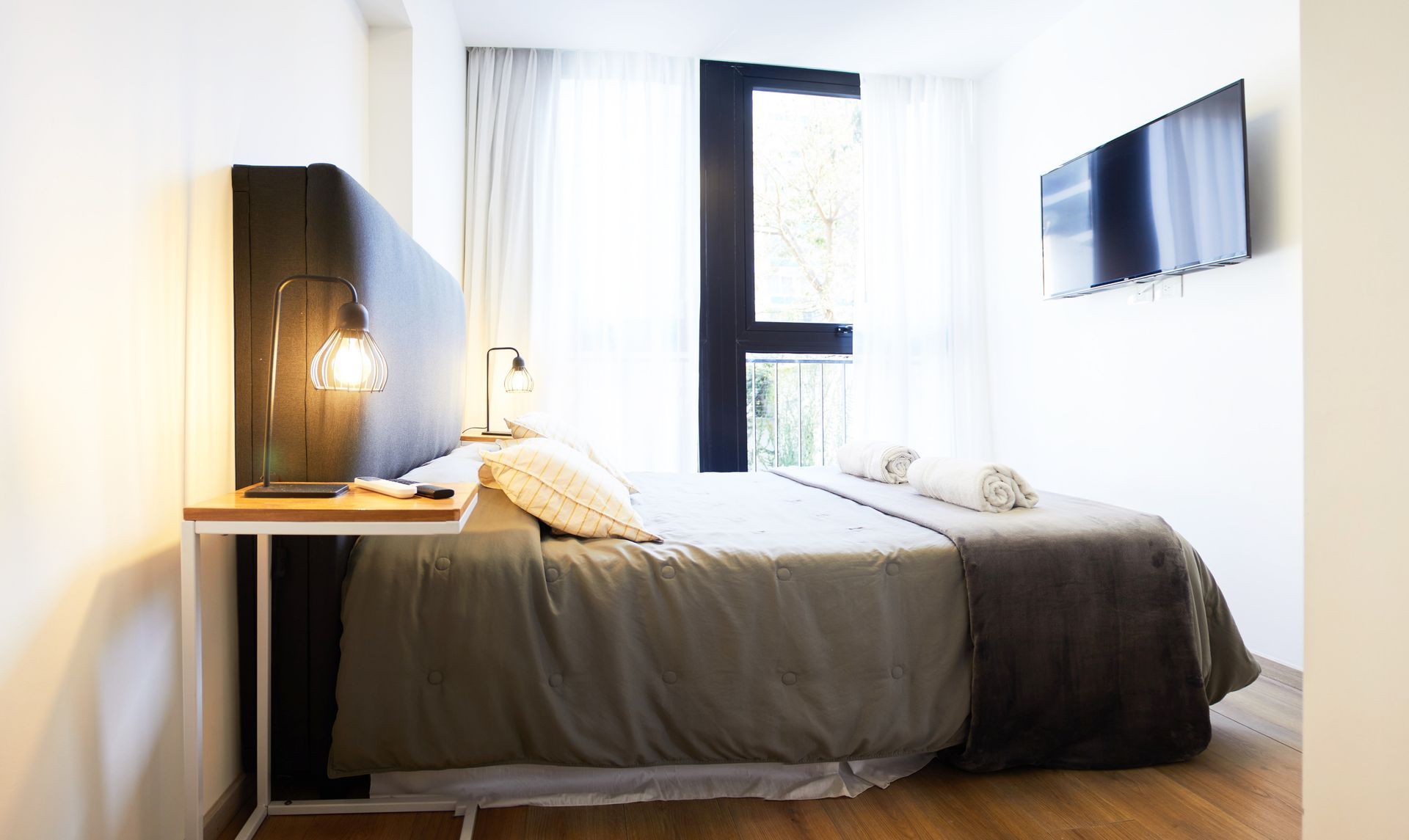 Dormitorio moderno con cama, lámpara de noche y televisor montado en la pared en una habitación luminosa y minimalista.