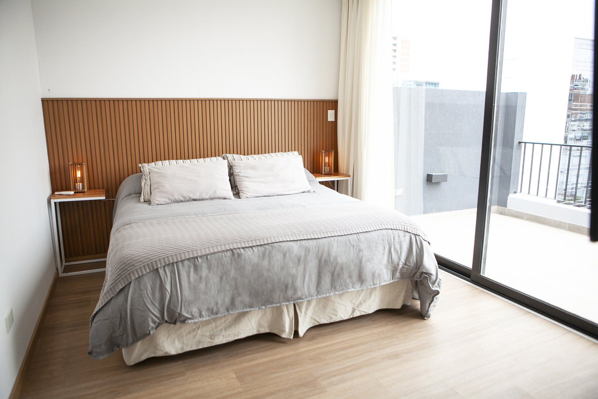 Habitación luminosa con cama gris, cabecero de madera, mesita de noche y puertas correderas de cristal que dan a un balcón.