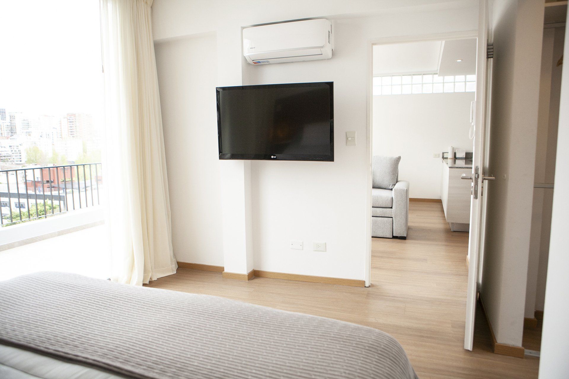 Habitación luminosa con cama, televisor de pared, aire acondicionado y puertas que dan a un balcón con vistas a la ciudad.