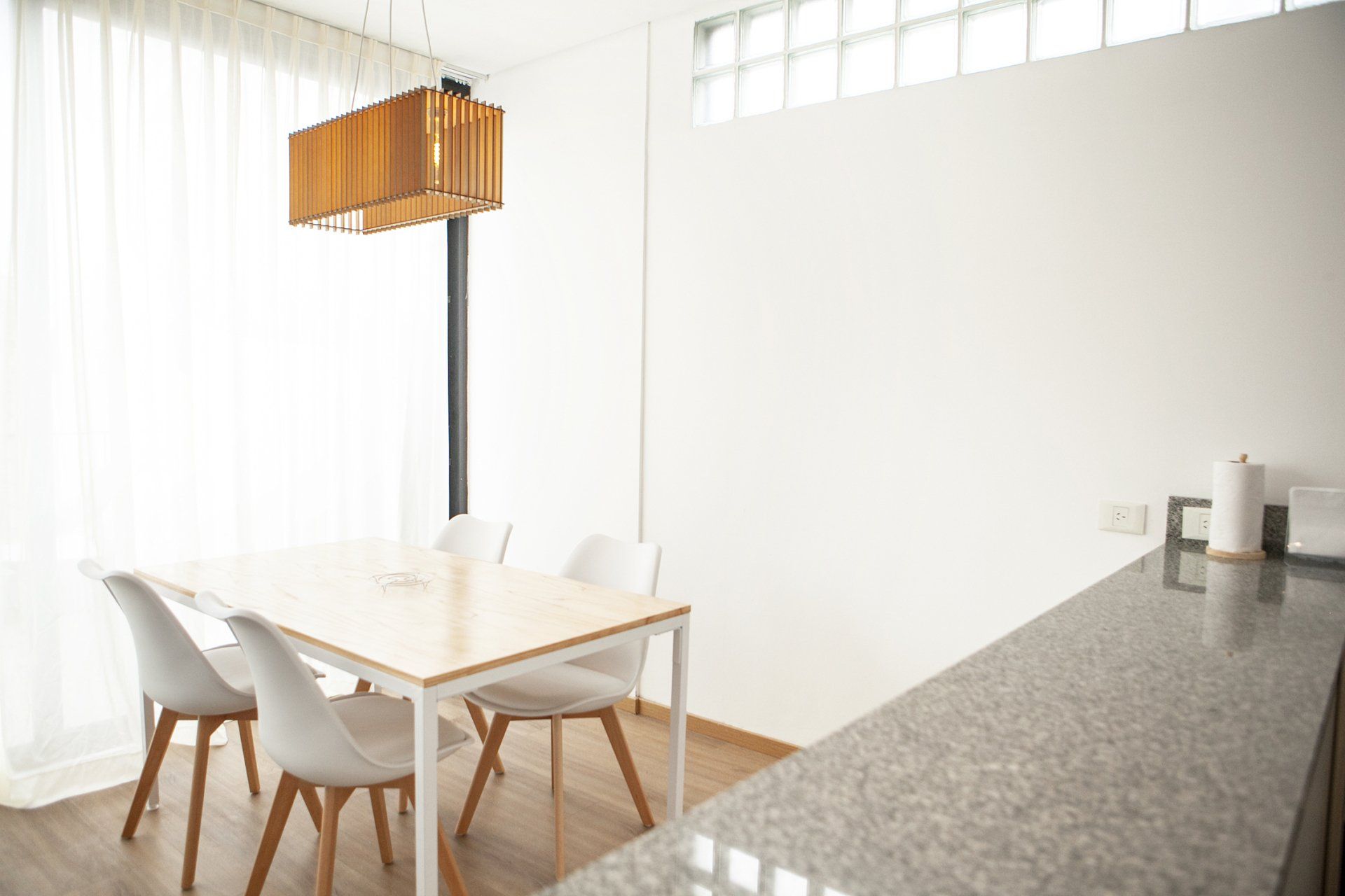 Comedor luminoso con sillas blancas, mesa de madera clara y una lámpara colgante de madera junto a la encimera de la cocina.