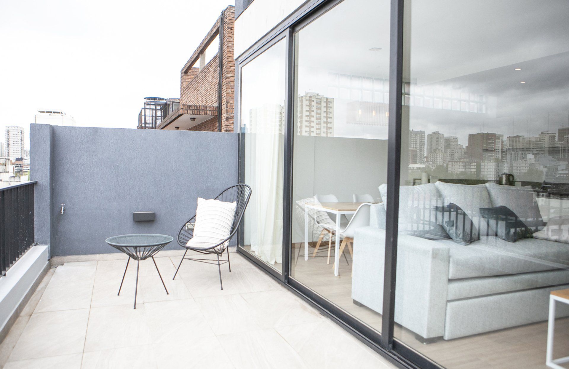 Balcón moderno con sillas de patio, una mesita y puertas de cristal junto a un luminoso salón.