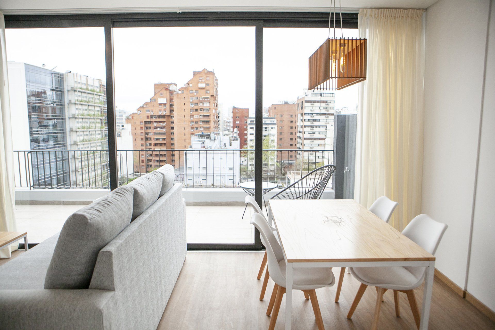 Sala de estar moderna con sofá gris, mesa de comedor blanca y grandes ventanales con vistas a los edificios de la ciudad.