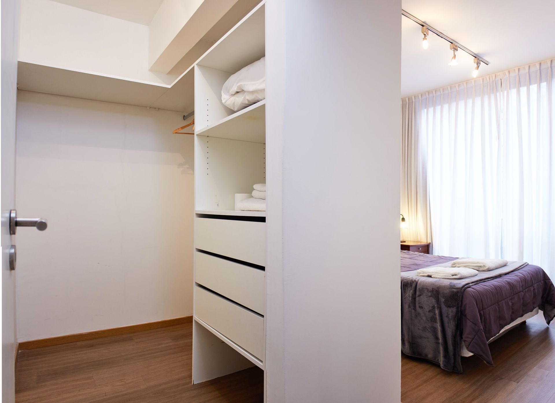 Armario abierto junto a un dormitorio con paredes blancas, suelo de madera y una cama junto a una ventana luminosa.