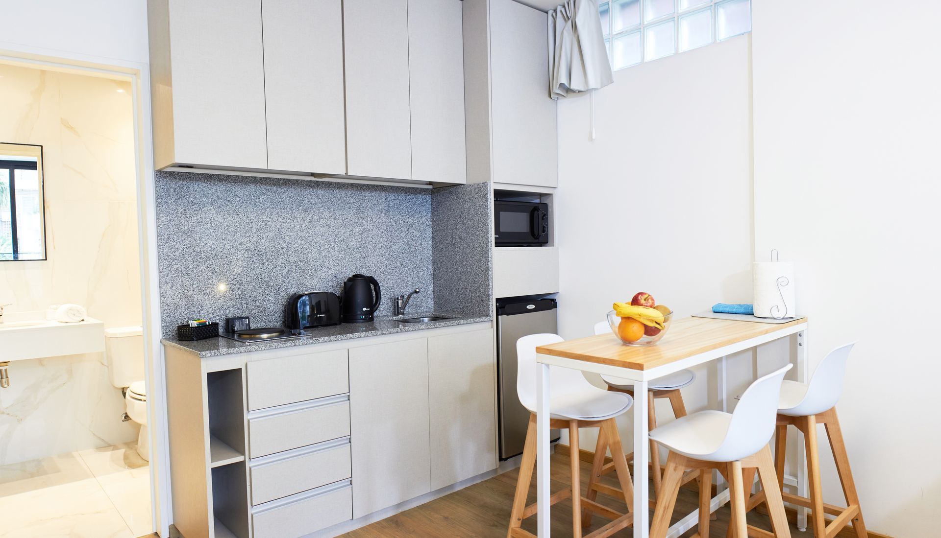 Cocina luminosa con armarios blancos, salpicadero de azulejos y una pequeña mesa de comedor con taburetes.