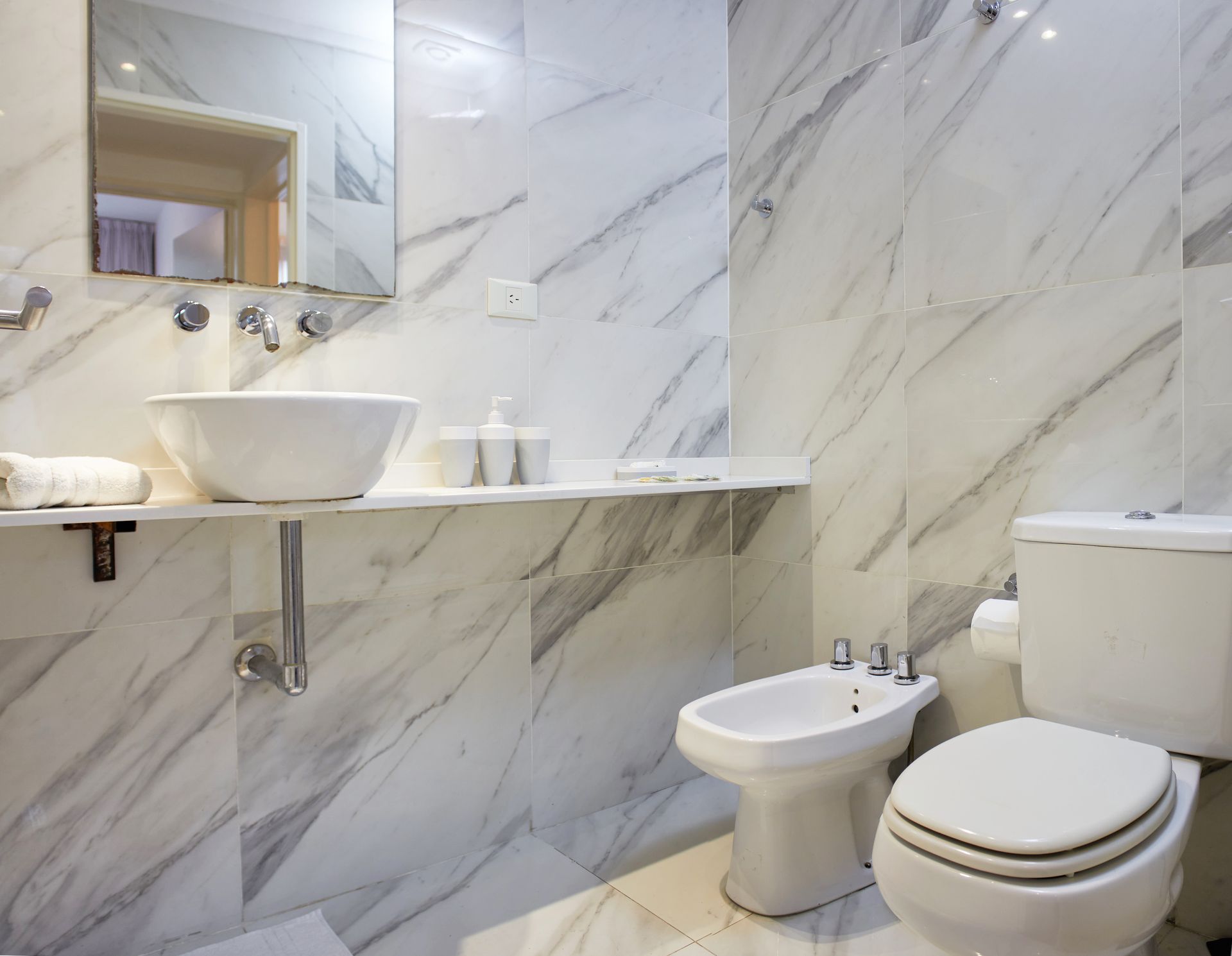 Baño moderno con paredes de mármol, lavabo, bidé e inodoro en tonos blancos.