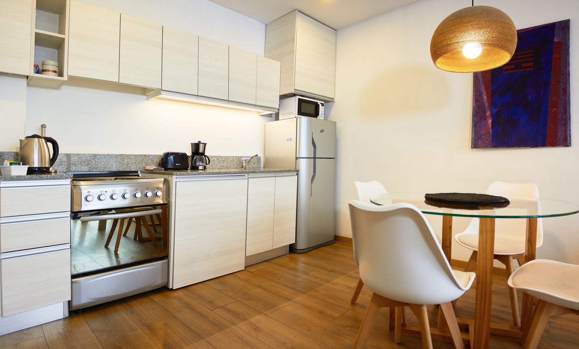 Cocina blanca y luminosa con estufa, refrigerador, mesa de comedor y piso de madera, iluminada por una lámpara colgante.