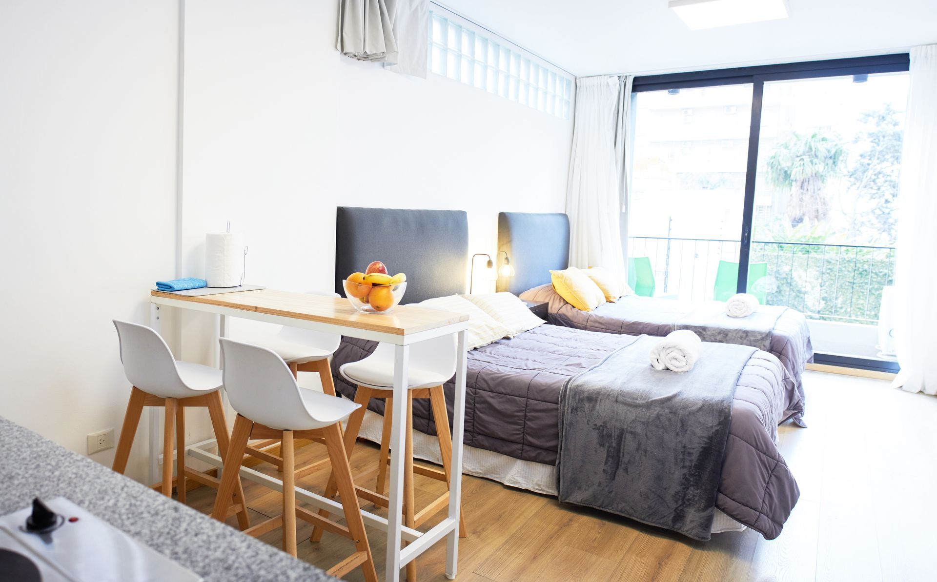 Habitación tipo estudio luminosa con cama, mesa de comedor y grandes puertas corredizas de vidrio que dan a un balcón.