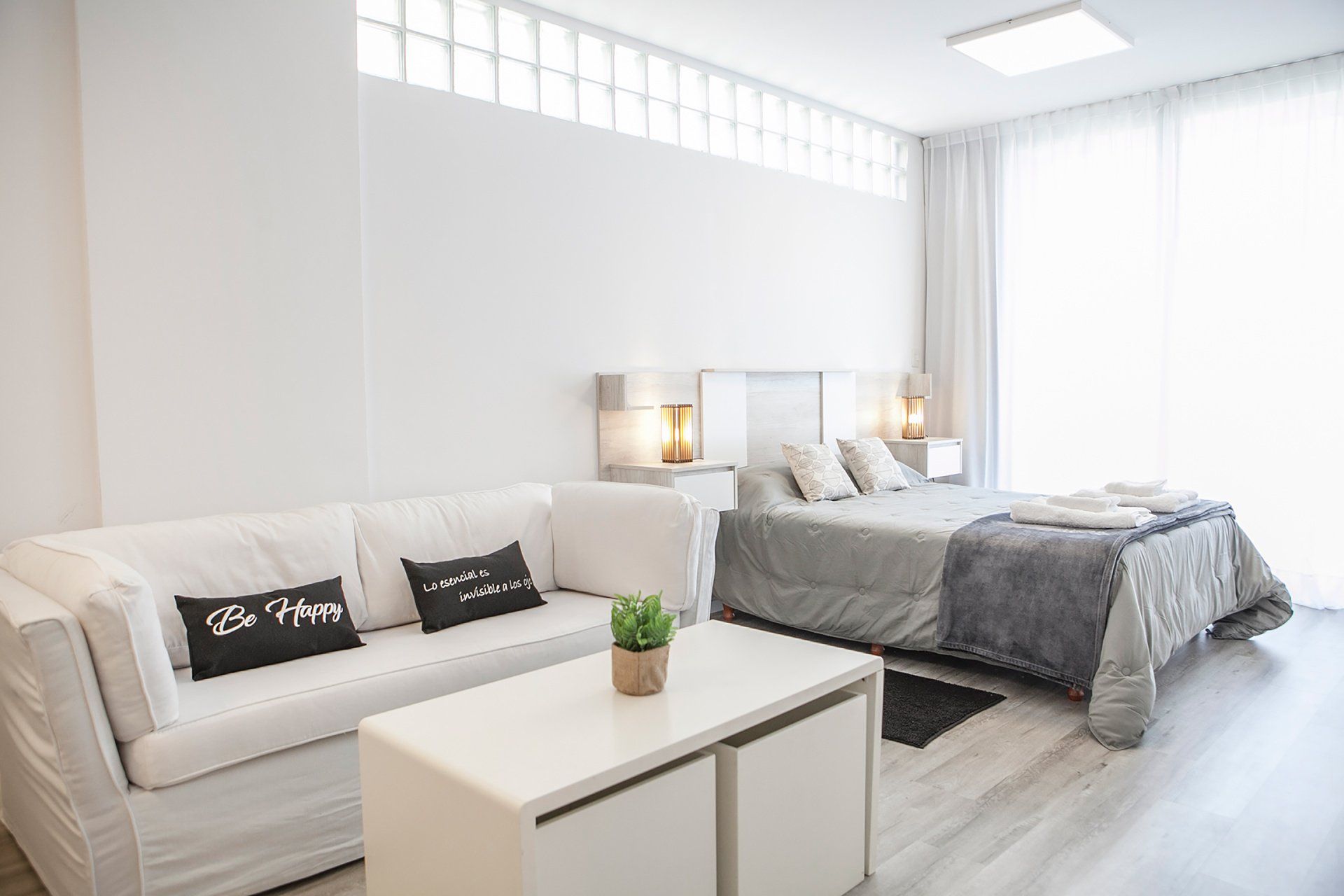 Dormitorio blanco luminoso con cama, sofá, escritorio y decoración en tonos grises suaves.