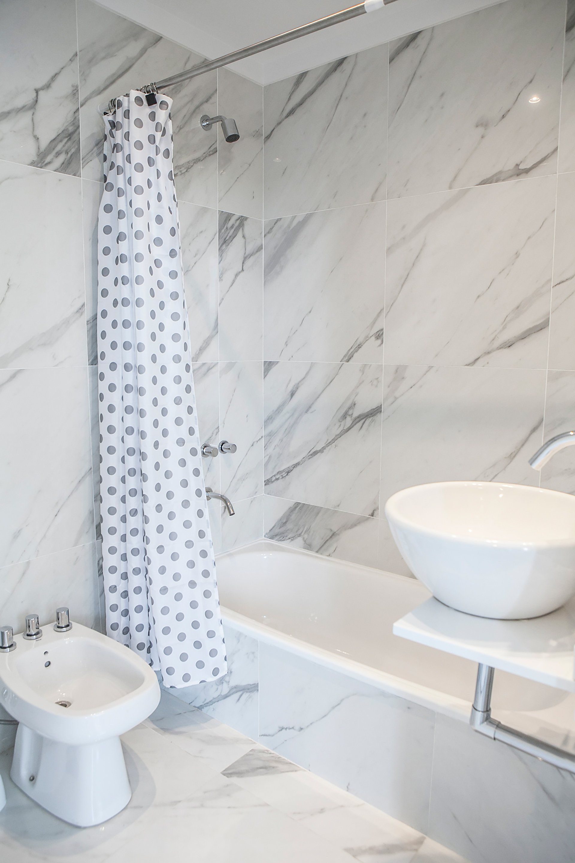 Baño moderno con paredes de mármol blanco, cortina de ducha, inodoro, bañera y lavabo redondo sobre encimera.