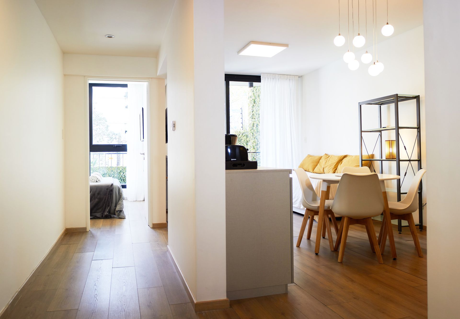 Pasillo luminoso junto a un comedor blanco con mesa de madera, sillas y lámparas colgantes en un apartamento moderno.