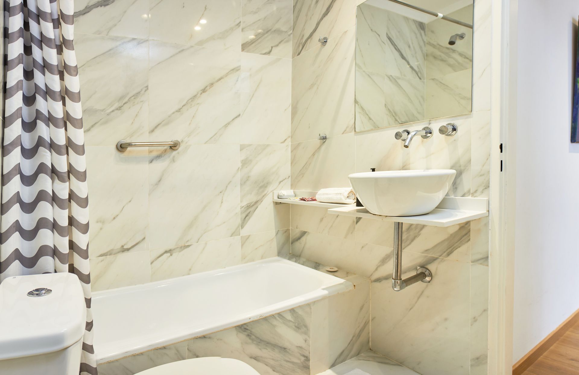 Baño de mármol con bañera, lavabo mural y cortina de ducha con estampado de espiga.