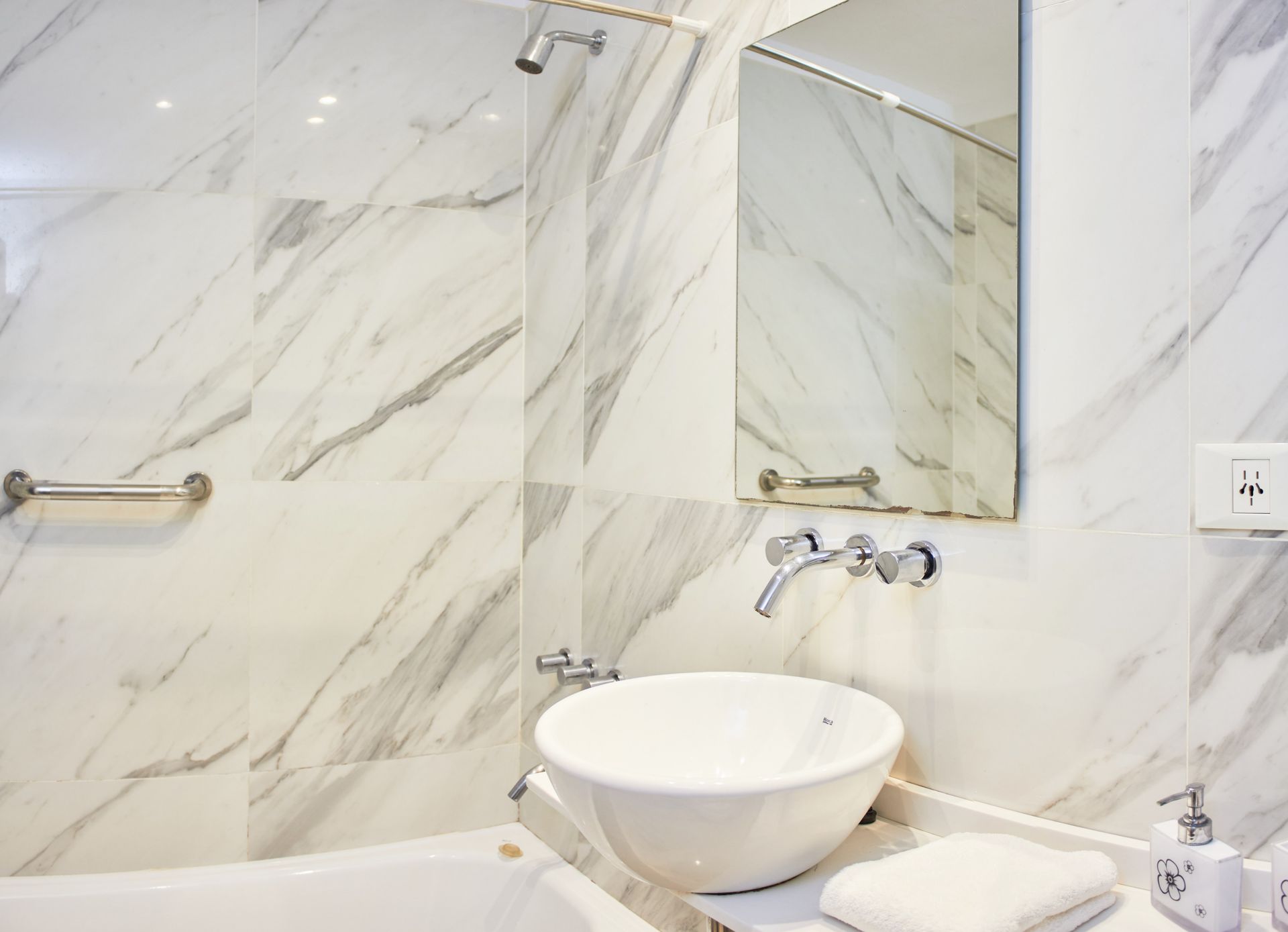 Baño moderno con azulejos de mármol, lavabo redondo blanco y espejo sobre la bañera.
