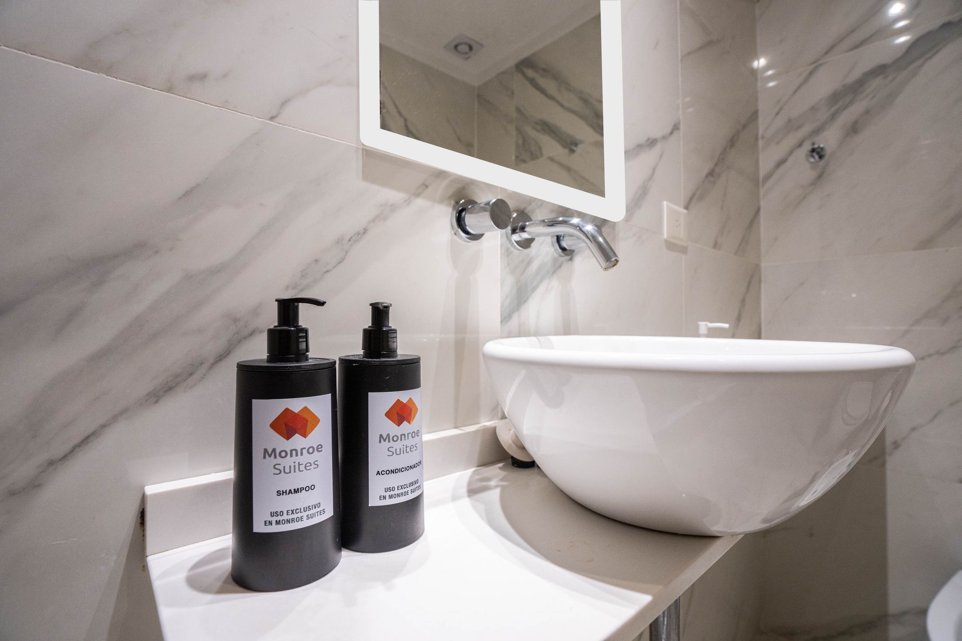 Tocador de baño moderno con lavabo blanco, paredes de mármol y dos dispensadores de jabón negros.