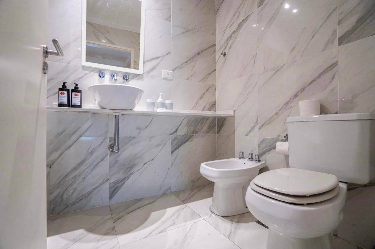 Baño moderno de color blanco con paredes de mármol, lavabo, espejo, bidé, inodoro y estante con artículos de aseo.