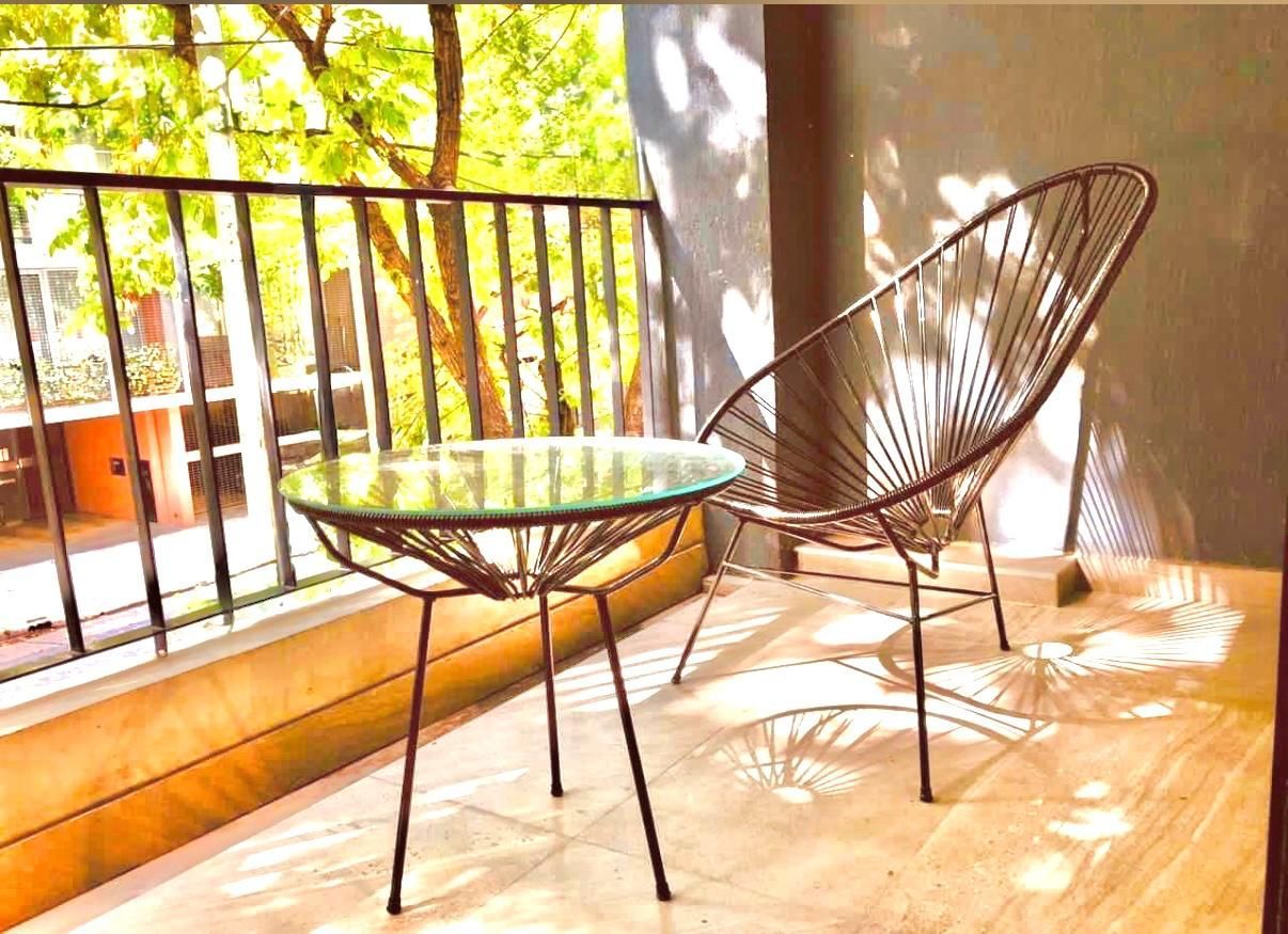 Balcón soleado con una mesa de cristal y una silla de alambre marrón sobre un suelo de madera.