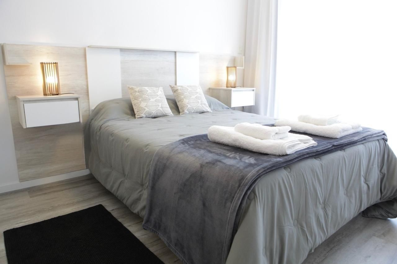 Dormitorio moderno con una colcha gris, almohadas blancas, lámparas de noche y toallas dobladas a los pies de la cama.