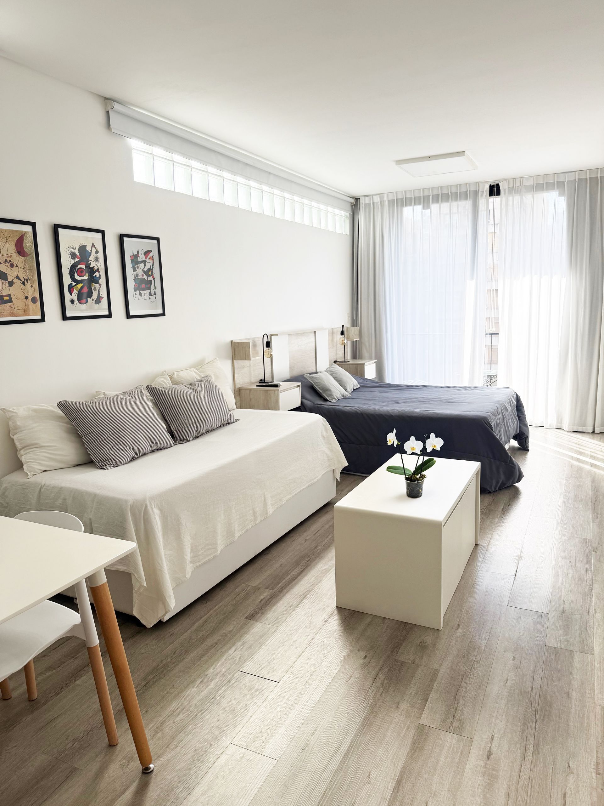 Dormitorio luminoso y minimalista con sofá blanco, cama, mesa de centro, cuadros enmarcados en la pared y cortinas transparentes.