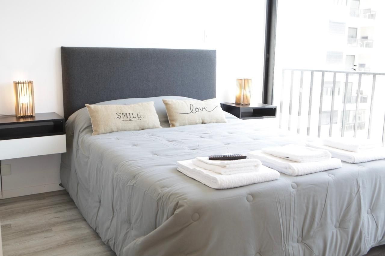 Habitación luminosa con cama gris, sábanas blancas, lámparas de noche y puerta que da al balcón.