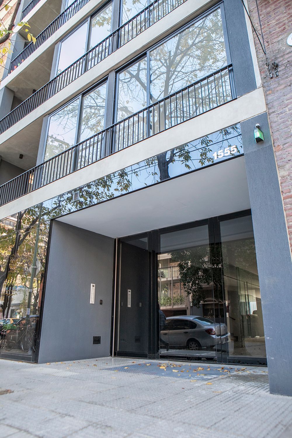 Entrada a un moderno edificio de apartamentos con puertas de cristal y balcones de metal negro, vista desde la acera.