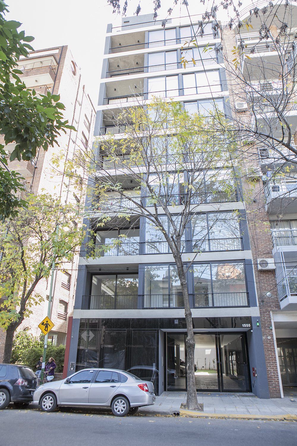 Edificio de apartamentos moderno de varias plantas con grandes ventanales y coches aparcados en una calle arbolada.