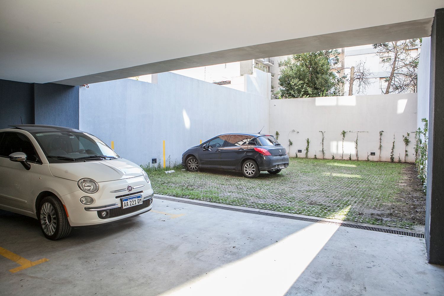 Entrada cubierta con dos coches aparcados junto a una pared blanca y un jardín con césped.
