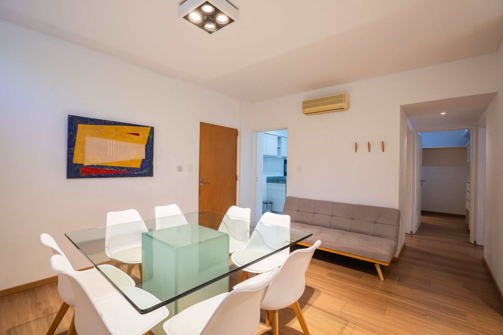 Luminoso salón-comedor con mesa de cristal, sillas blancas, sofá, cuadros en la pared y pasillo abierto.