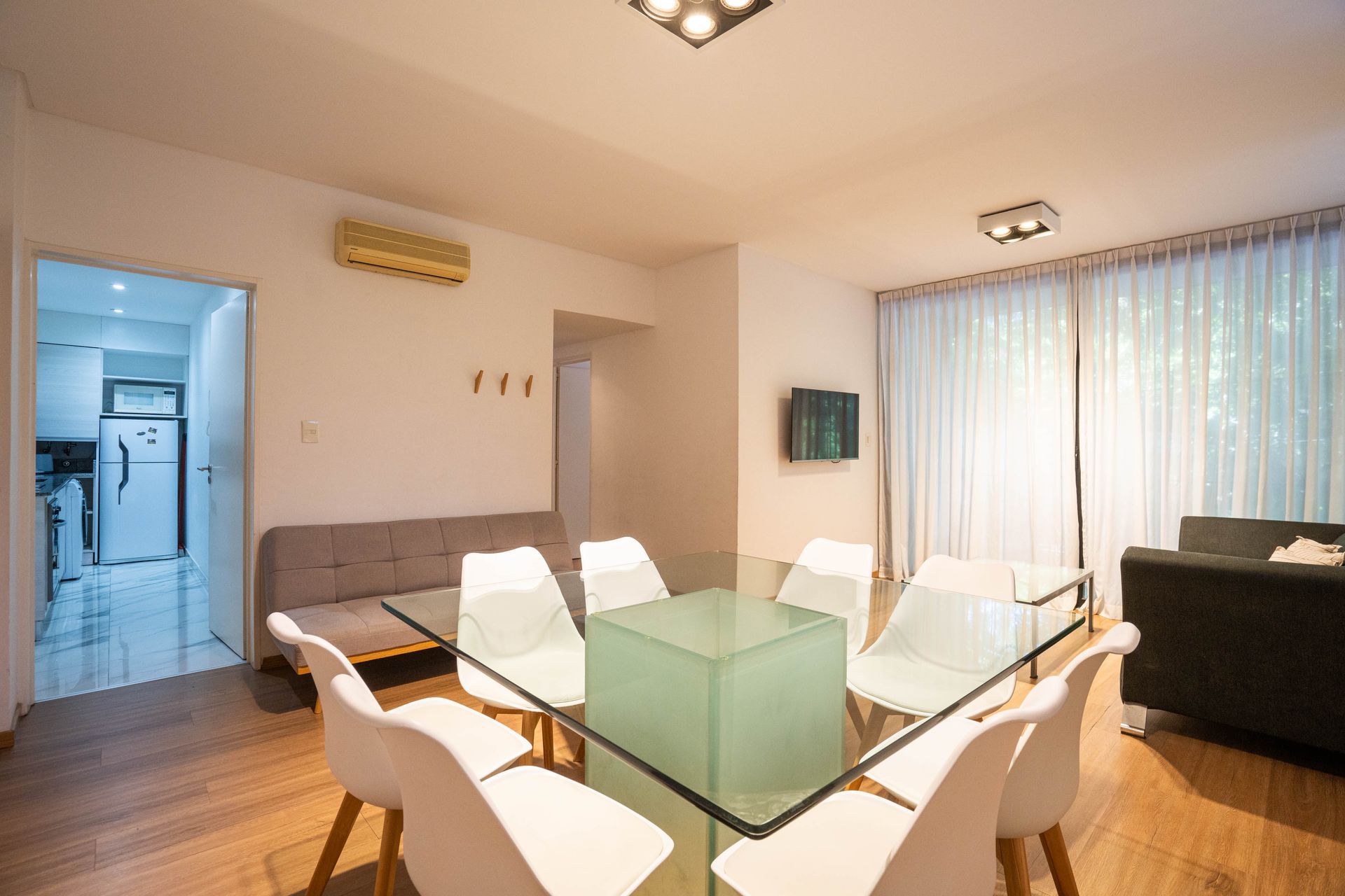 Comedor moderno con mesa de cristal, sillas blancas, sofá gris y grandes cortinas en las ventanas.