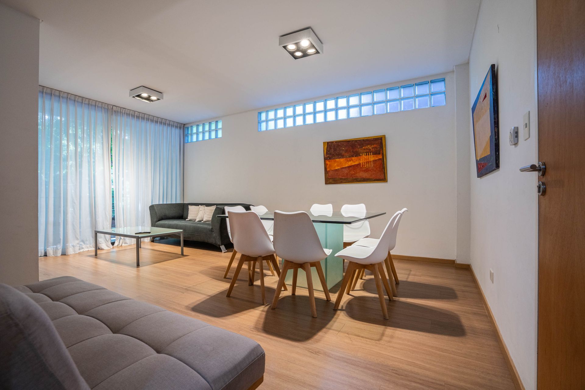 Comedor moderno con mesa de cristal, sillas blancas, sofá y grandes ventanales.