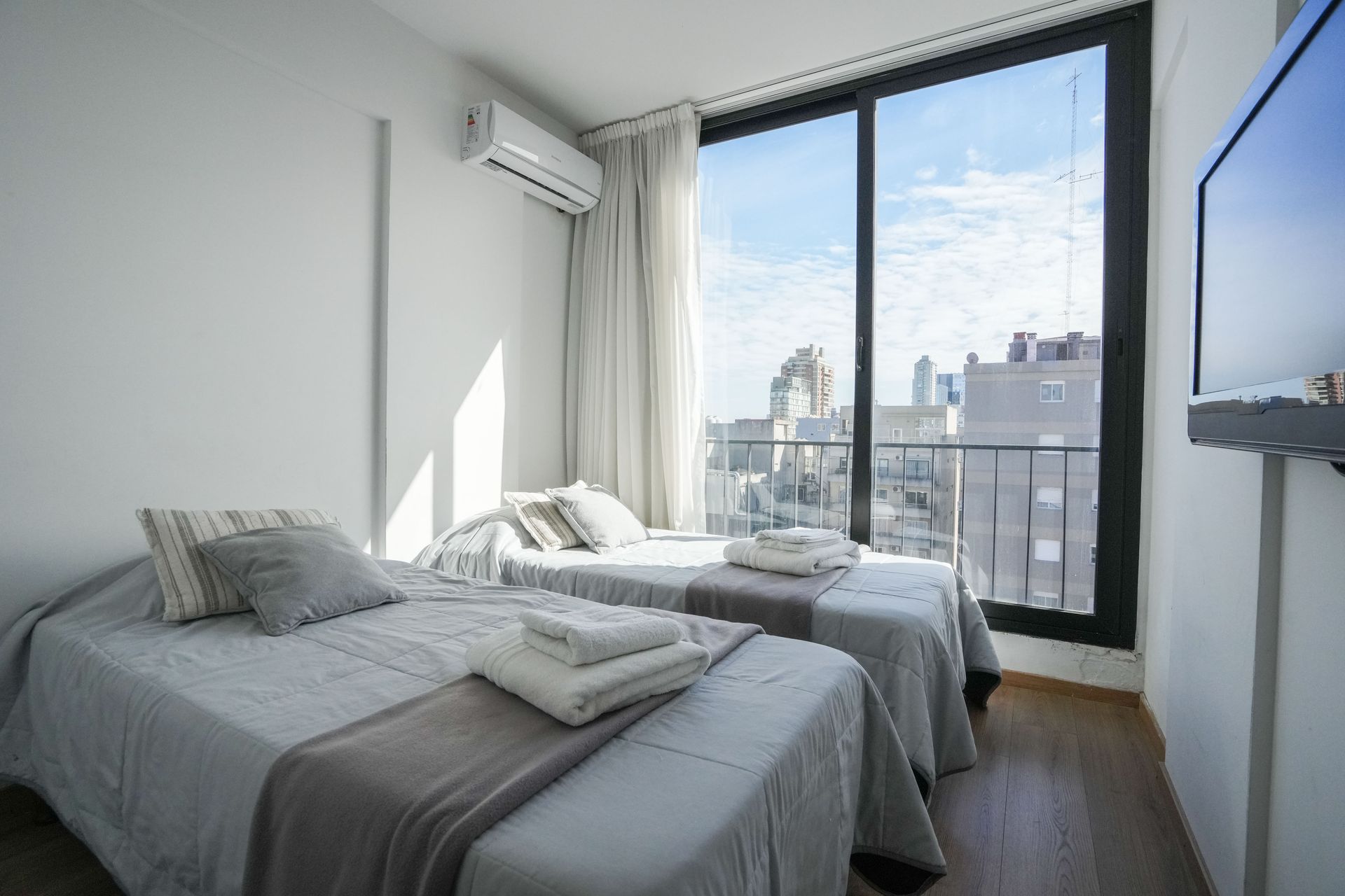 Habitación luminosa con dos camas grises, ventana grande, cortinas y televisor montado en la pared.