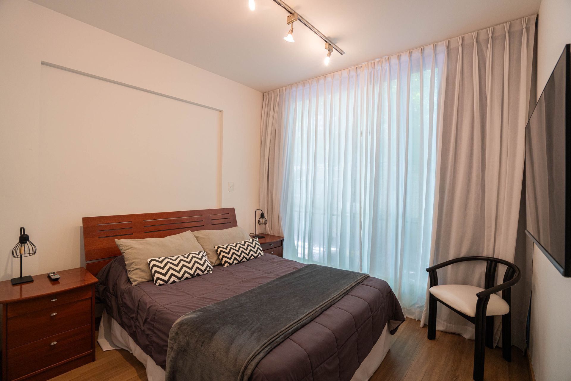 Dormitorio moderno con cama, mesita de noche, silla y cortinas que van del suelo al techo junto a una gran ventana.
