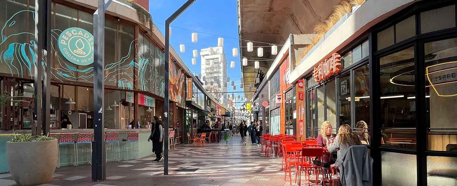 Calle peatonal comercial con mesas de café al aire libre, escaparates de cristal y edificios altos a lo lejos.